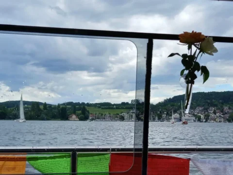 Blick durch ein Fenster auf einen See, Segelboote, grüne Hügel und Gebetsfahnen am Fenster.