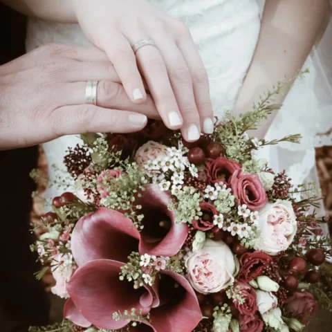 Hände eines Brautpaars mit Eheringen über einem Blumenstrauß halten.