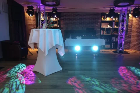 Stehtisch mit weißer Tischdecke auf beleuchteter Tanzfläche in gemütlicher Bar mit Backsteinwänden.