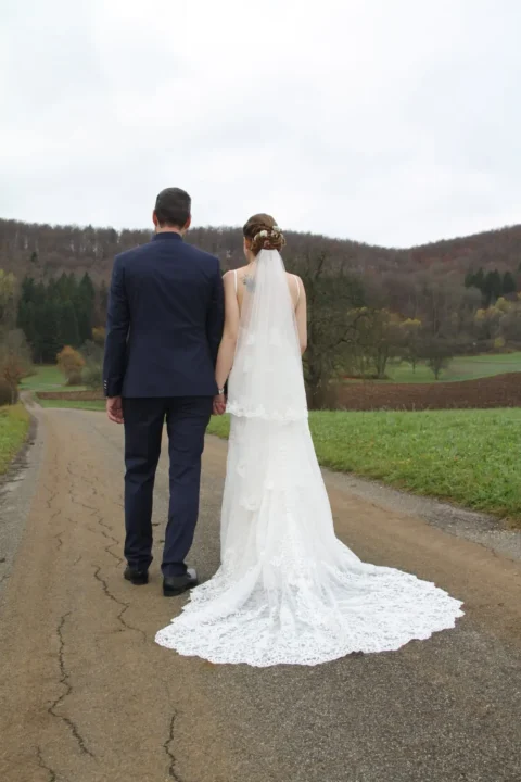 Ein Hochzeitspaar in Hochzeitskleidung, Hand in Hand, geht eine Landstraße entlang in Richtung eines bewaldeten Hügels.