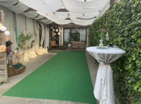 Überdachter Eingangsbereich zum Feiern, mit grüner Kunstrasen, Stehtisch und dekorativen Pflanzen.