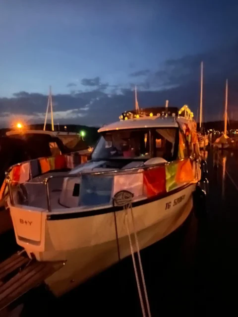 Boot im Hafen bei Nacht, beleuchtet mit bunten Lichterketten und umgeben von anderen Booten unter einem dunklen Himmel.