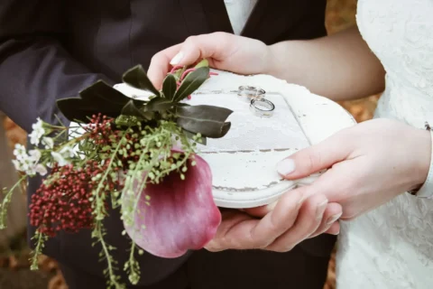 Braut und Bräutigam halten Blumenstrauß mit Eheringen auf weißem Tablett am Hochzeitstag.