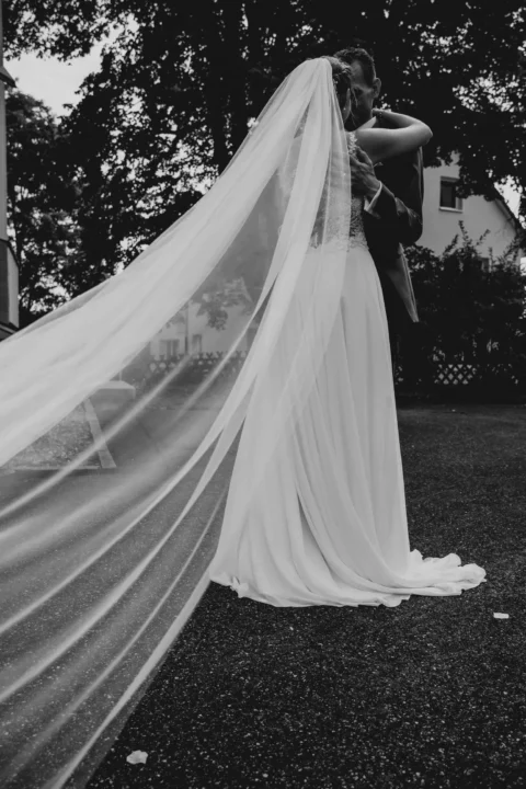 Brautpaar umarmt sich innig im Freien, langer Schleier weht romantisch im Wind, schwarz-weiß Foto.