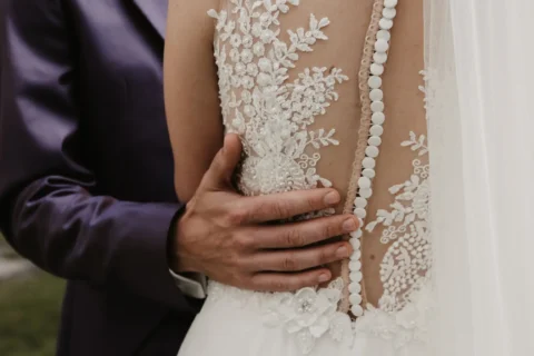 Bräutigam umarmt Braut in Spitzen-Hochzeitskleid mit Knöpfen am Rücken.