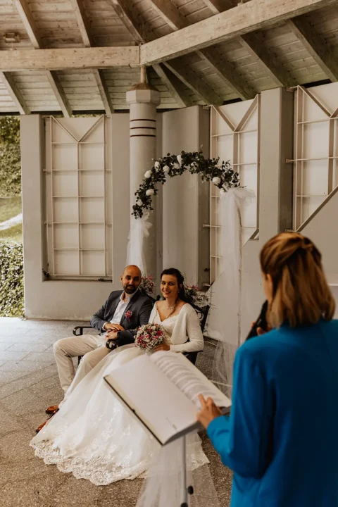 Traurednerin hält emotionale Zeremonie für Brautpaar unter romantischem Blütenbogen ab.