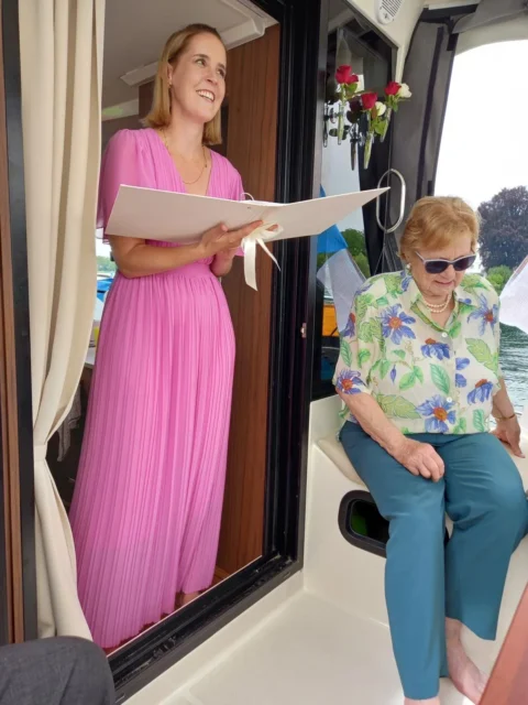 Traurednerin hält feierliche Rede auf Boot, begleitet von älterer Dame, in entspannter Atmosphäre.