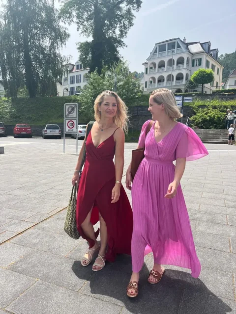 Zwei Frauen in eleganten Sommerkleidern spazieren lächelnd in sonniger Umgebung.
