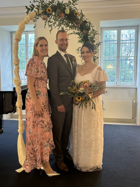 Ein Hochzeitspaar mit Traurednerin bei der Zeremonie, vor einem Blumenbogen stehend, Traurednerin in rosa Kleid.