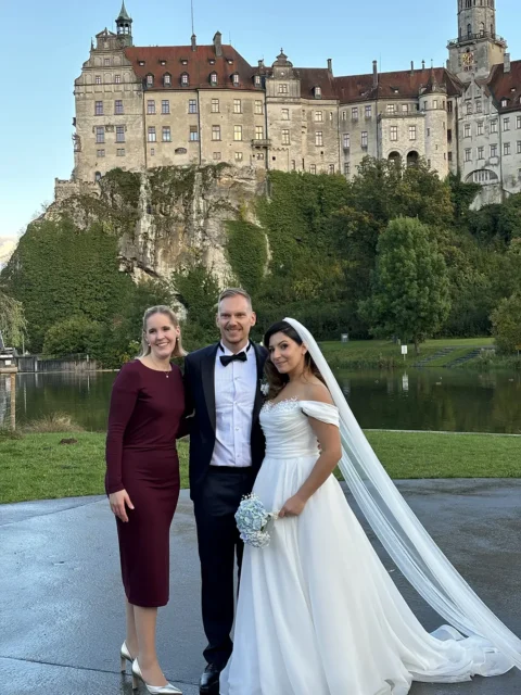 Brautpaar mit Traurednerin vor einem historischen Schloss im Grünen, romantische Hochzeit im Freien.