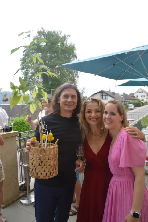 Drei Personen bei einer Feier im Freien mit einem Weidenkorb und Gläsern; fröhliche Stimmung.