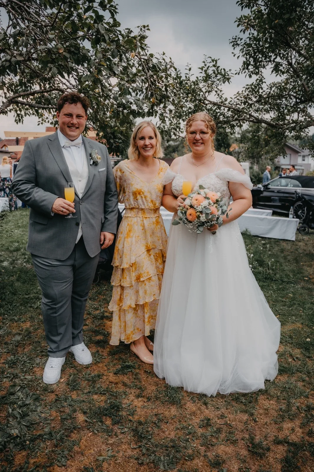Hochzeitsfeier mit Braut und Gästen im Freien, alle lächeln und halten Getränke in der Hand. Die Braut trägt ein elegantes, weißes Kleid und hält einen Blumenstrauß.