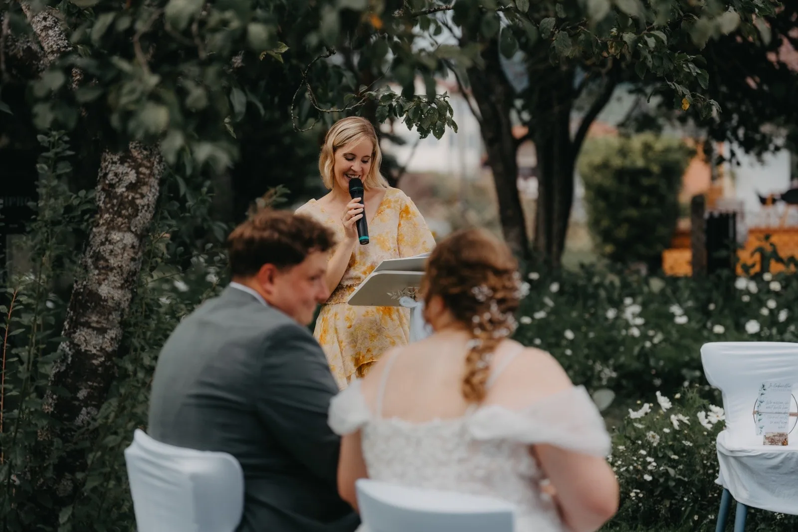 Hochzeitszeremonie im Freien mit dem Brautpaar, das den Redner aufmerksam zuhört, umgeben von blühenden Pflanzen.