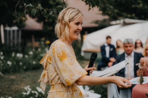 Frau in einem blumigen Kleid hält ein Mikrofon und spricht bei einer Outdoor-Veranstaltung, umgeben von Gästen in festlicher Kleidung und einer schönen Gartenlandschaft.