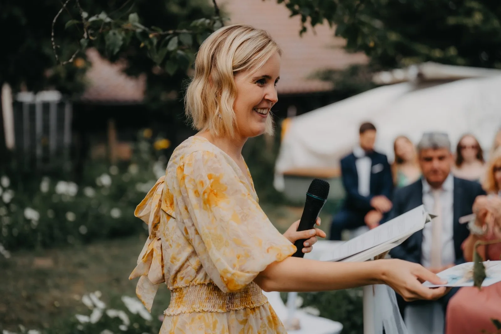 Frau in einem blumigen Kleid hält ein Mikrofon und spricht bei einer Outdoor-Veranstaltung, umgeben von Gästen in festlicher Kleidung und einer schönen Gartenlandschaft.