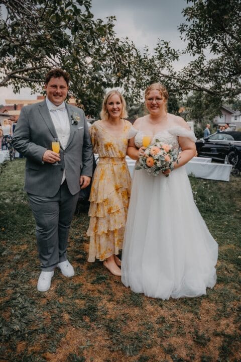 Hochzeitsfeier mit Braut und zwei Gästen, die in eleganter Kleidung posieren. Die Braut trägt ein weißes Kleid und hält einen Blumenstrauß, während die anderen beiden Gläser mit Getränken in der Hand halten. Im Hintergrund sind Tische und eine Gartenlandschaft sichtbar.