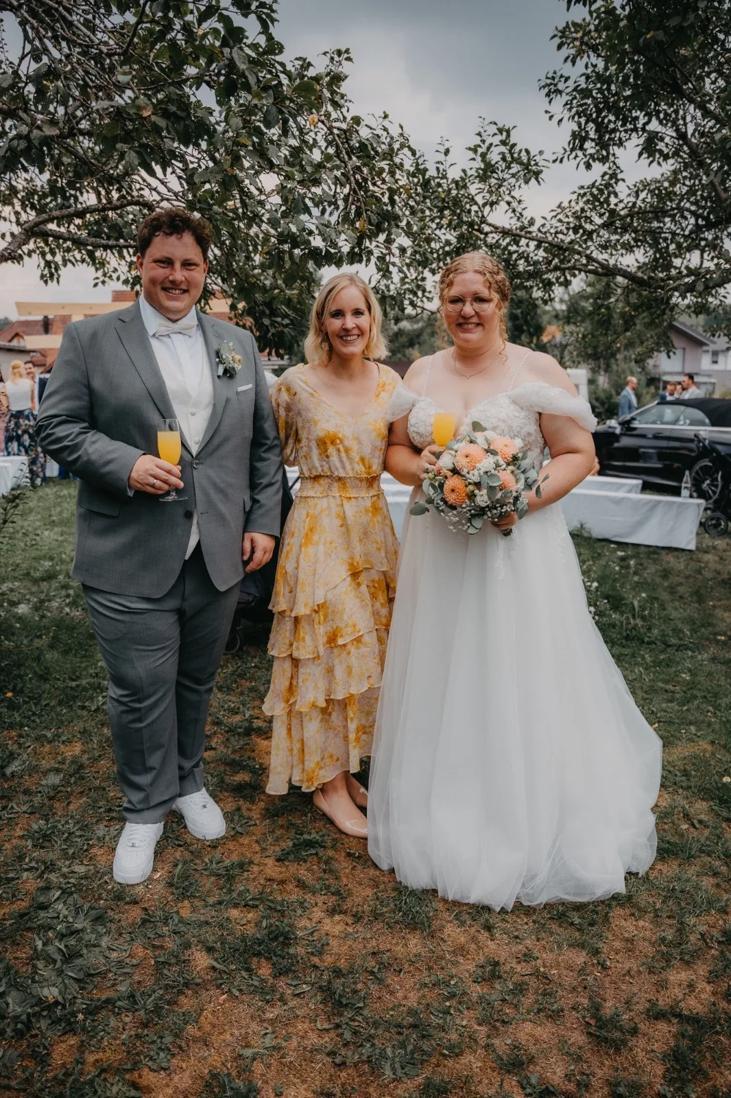 Hochzeitsfeier mit Braut und zwei Gästen, die in eleganter Kleidung posieren. Die Braut trägt ein weißes Kleid und hält einen Blumenstrauß, während die anderen beiden Gläser mit Getränken in der Hand halten. Im Hintergrund sind Tische und eine Gartenlandschaft sichtbar.