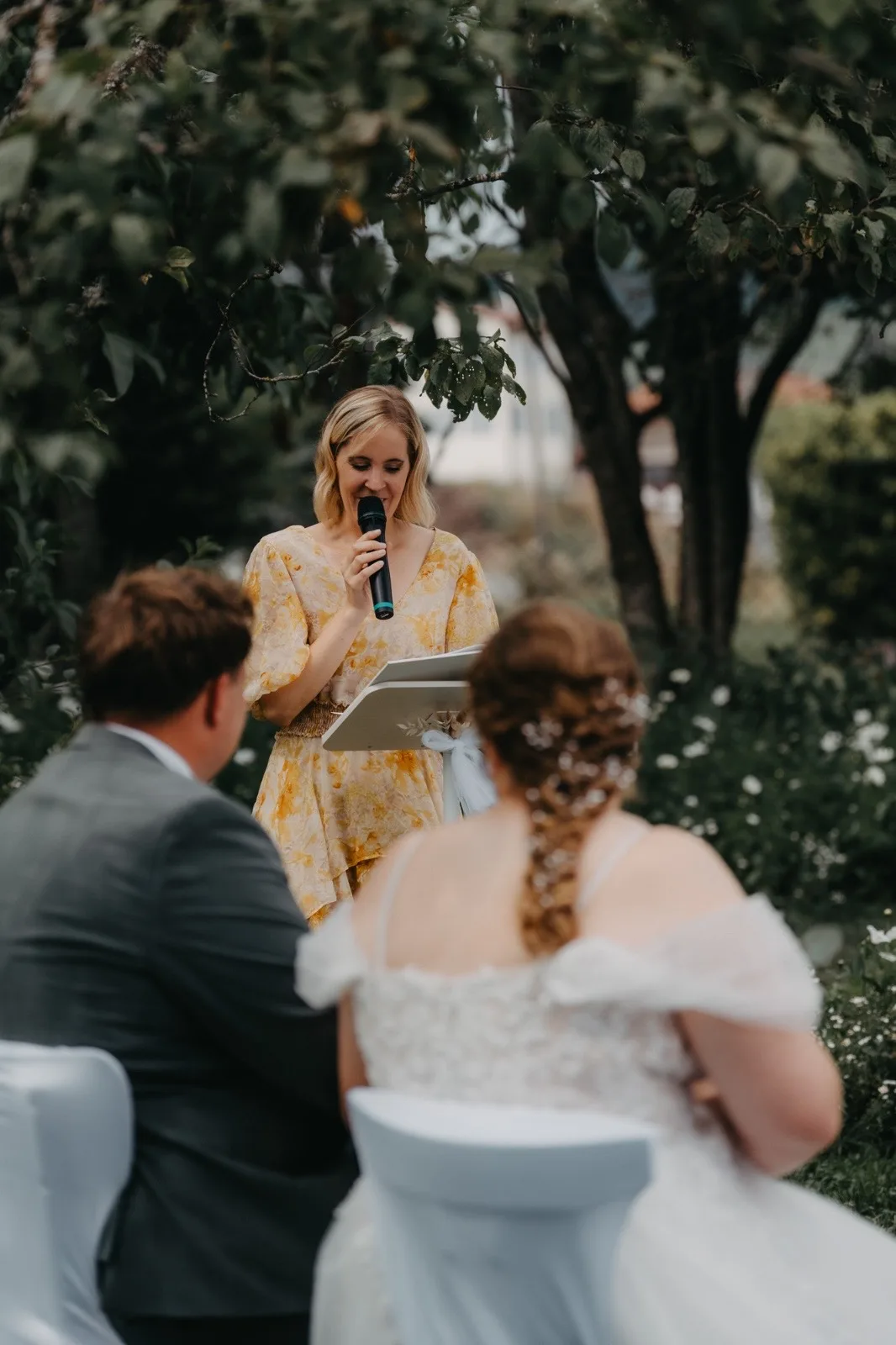 Hochzeitszeremonie im Freien, mit einer Rednerin in einem gelben Kleid, die das Brautpaar anspricht. Im Hintergrund blühende Pflanzen und eine romantische Atmosphäre.