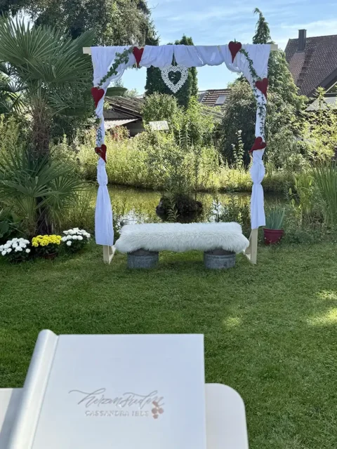 Ein Hochzeitsdekor im Freien mit einem mit weißen Stoffen und roten Herzen geschmückten Bogen, vor einem kleinen Teich. Ein mit Fell bezogener Sitz steht darunter, im Vordergrund ein Buch auf einem Tisch mit verziertem Umschlag.