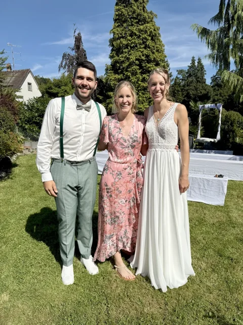 Drei Menschen stehen lächelnd im Freien auf einer grünen Wiese: ein Mann in weißem Hemd und Hosenträgern, eine Frau in einem blumigen Kleid und eine Frau in einem weißen Kleid.