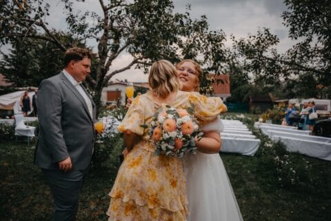 Hochzeitsfeier im Freien mit zwei Frauen, die sich umarmen. Eine Braut in einem weißen Kleid hält einen Blumenstrauß, während die andere Frau in einem gelben Kleid mit Blumenmuster strahlt. Im Hintergrund sind Hochzeitsstühle und Bäume sichtbar.