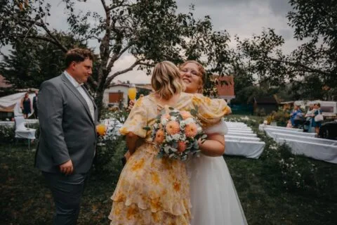 Hochzeitsfeier im Freien mit zwei Frauen, die sich umarmen. Eine Braut in einem weißen Kleid hält einen Blumenstrauß, während die andere Frau in einem gelben Kleid mit Blumenmuster strahlt. Im Hintergrund sind Hochzeitsstühle und Bäume sichtbar.
