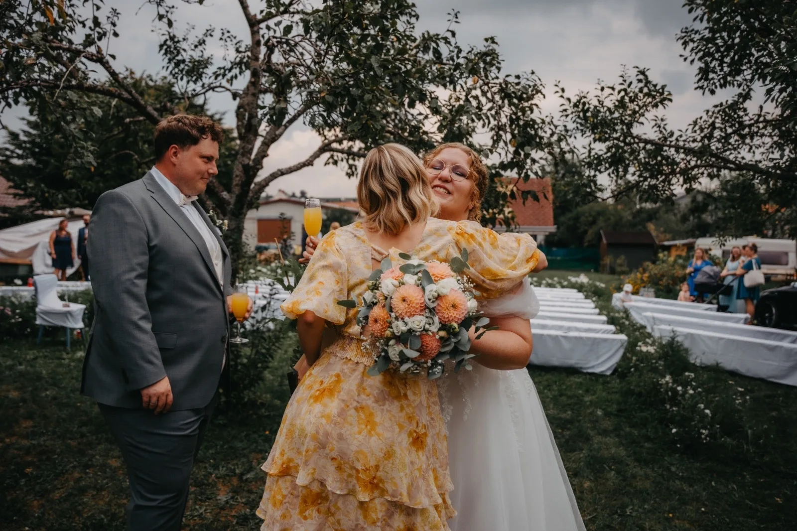 Hochzeitsfeier im Freien mit zwei Frauen, die sich umarmen. Eine Braut in einem weißen Kleid hält einen Blumenstrauß, während die andere Frau in einem gelben Kleid mit Blumenmuster strahlt. Im Hintergrund sind Hochzeitsstühle und Bäume sichtbar.