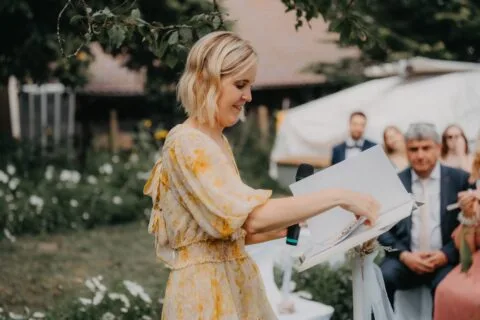 Eine Frau in einem blumigen Kleid hält ein Notizbuch und spricht bei einer Hochzeitszeremonie im Freien, umgeben von Blumen und Gästen.