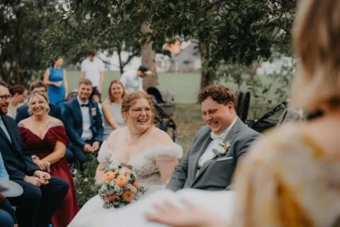 Braut und Bräutigam lächeln während ihrer Zeremonie im Freien, umgeben von Gästen in festlicher Kleidung und einer romantischen Atmosphäre.