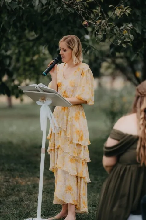 Frau in einem gelben, blumigen Kleid spricht mit einem Mikrofon während einer Zeremonie im Freien, im Hintergrund sind Bäume zu sehen.