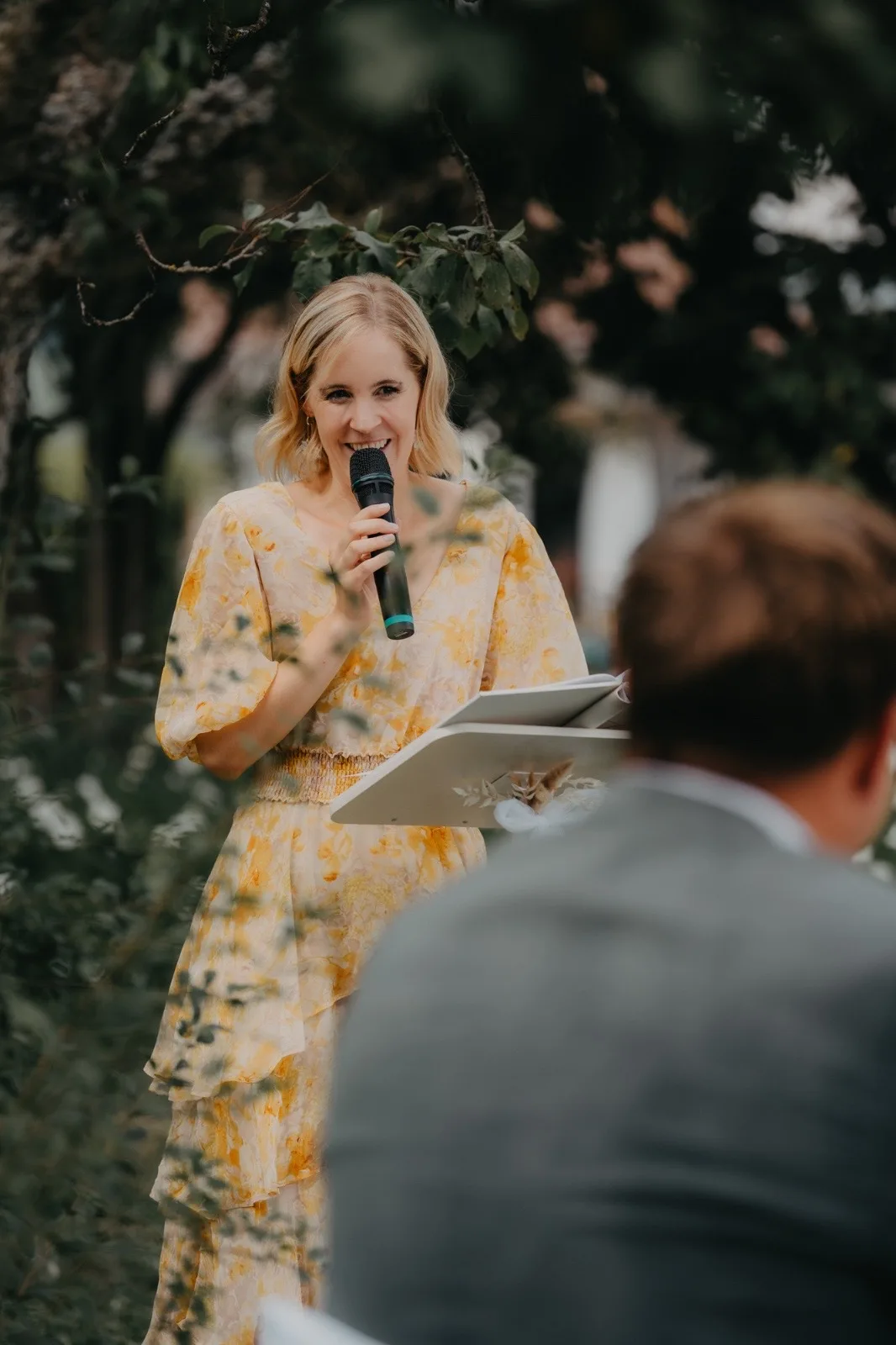 Eine Frau in einem blumigen gelben Kleid spricht mit einem Mikrofon bei einer Zeremonie im Freien, während Gäste im Hintergrund zuhören.