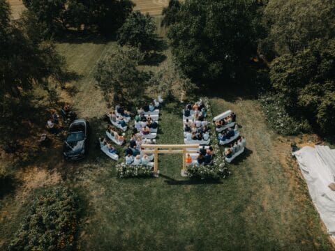 Hochzeitszeremonie im Freien mit Gästen, die auf Bänken sitzen. Eine Holzstruktur als Hintergrund, umgeben von Bäumen und Blumen. Ein Auto steht in der Nähe, während die Atmosphäre festlich und entspannt wirkt.
