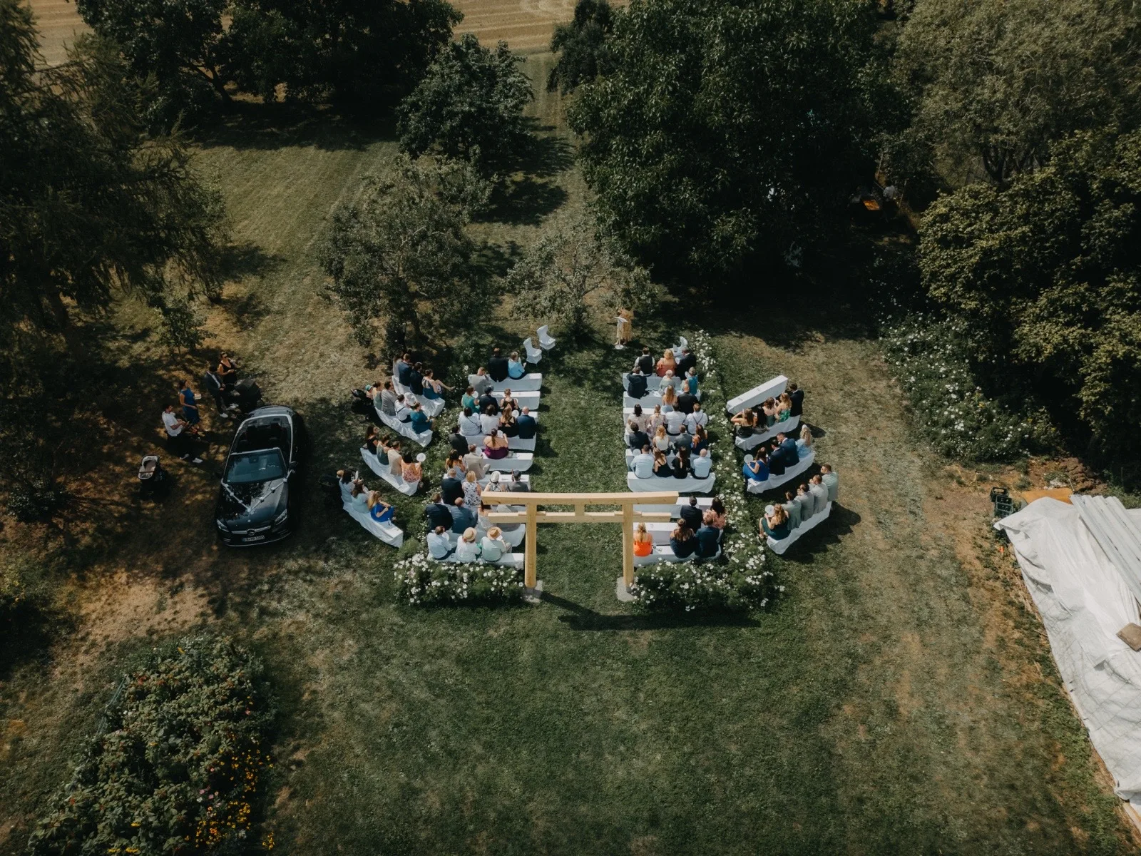 Hochzeitszeremonie im Freien mit Gästen, die auf Bänken sitzen. Eine Holzstruktur als Hintergrund, umgeben von Bäumen und Blumen. Ein Auto steht in der Nähe, während die Atmosphäre festlich und entspannt wirkt.